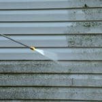 A pressure washer removing mold and biological growth from vinyl siding on a home exterior.
