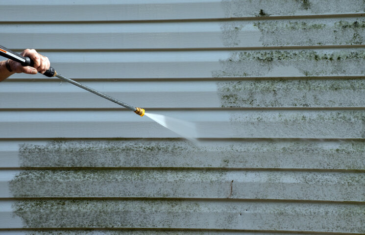 A pressure washer removing mold and biological growth from vinyl siding on a home exterior.