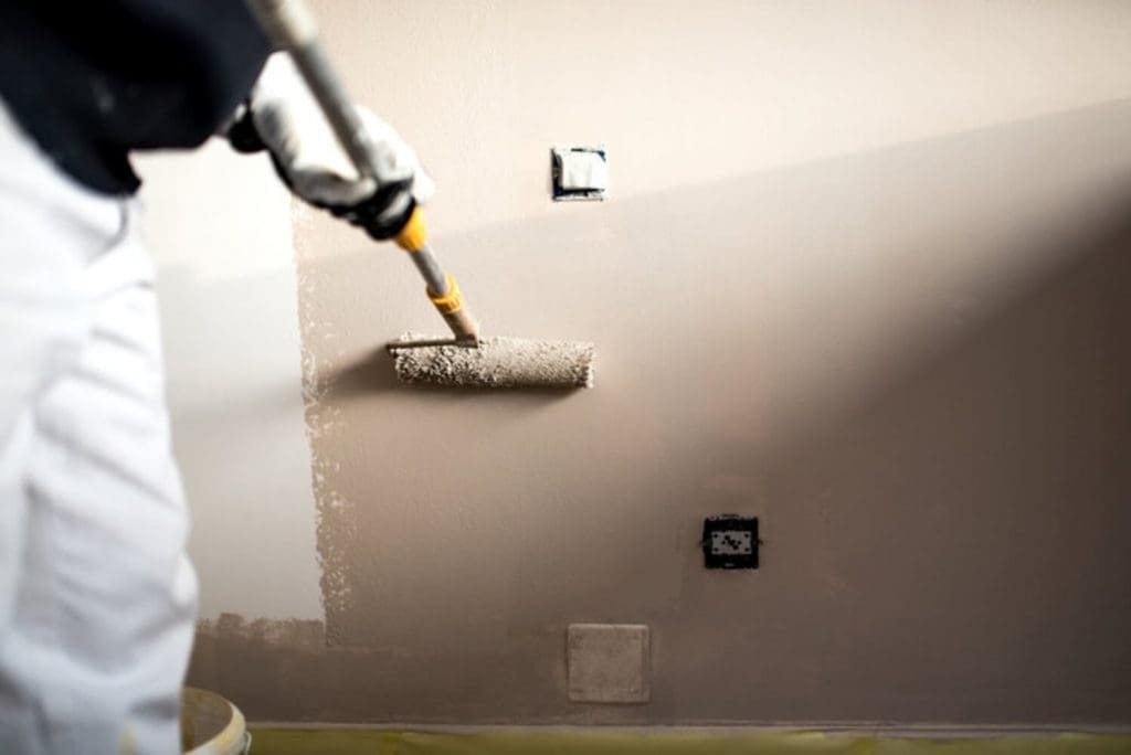 A painter uses a roller to apply paint to an interior wall during a professional interior painting project, with outlets taped off for protection.