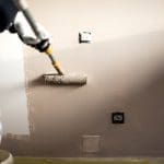 A painter uses a roller to apply paint to an interior wall during a professional interior painting project, with outlets taped off for protection.