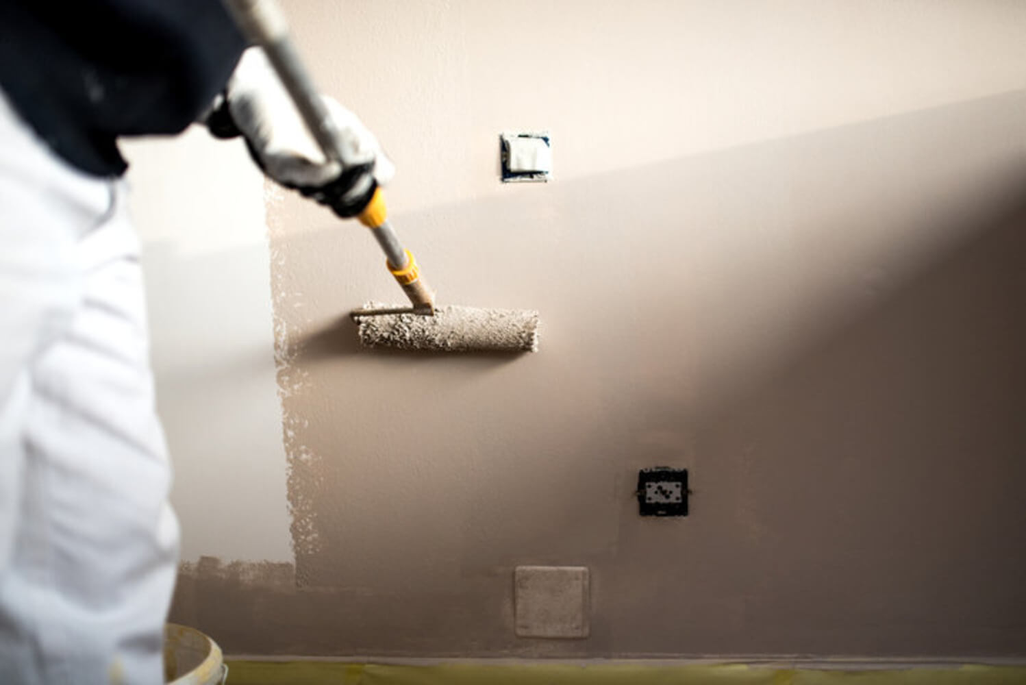A painter uses a roller to apply paint to an interior wall during a professional interior painting project, with outlets taped off for protection.