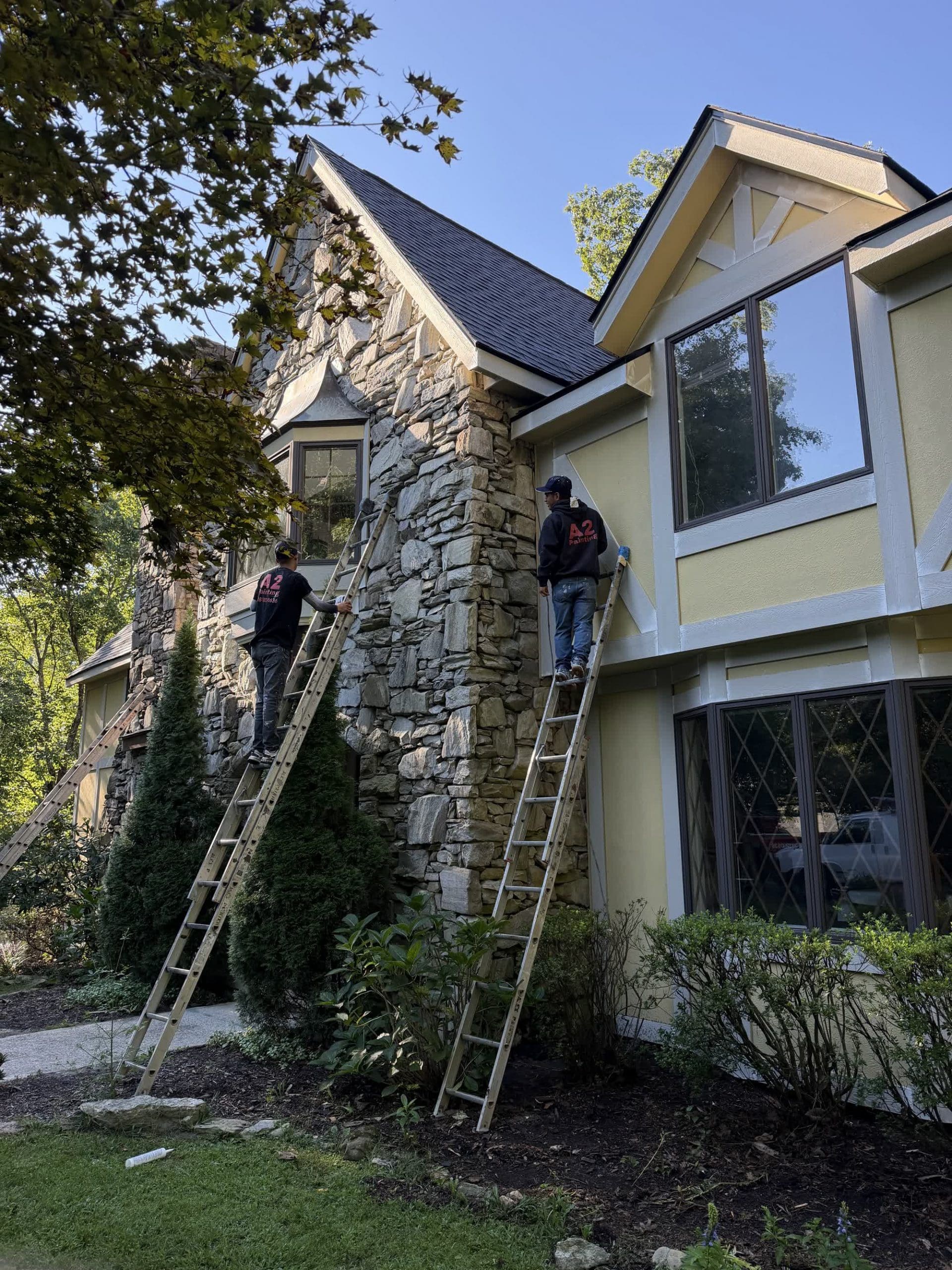 Professional painters working on extension ladders to prep and paint the natural stone exterior of a home in Hendersonville, NC