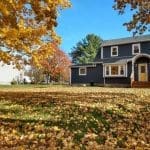 A two-story home with dark blue siding surrounded by fall foliage, illustrating the seasonal window for exterior house painting.