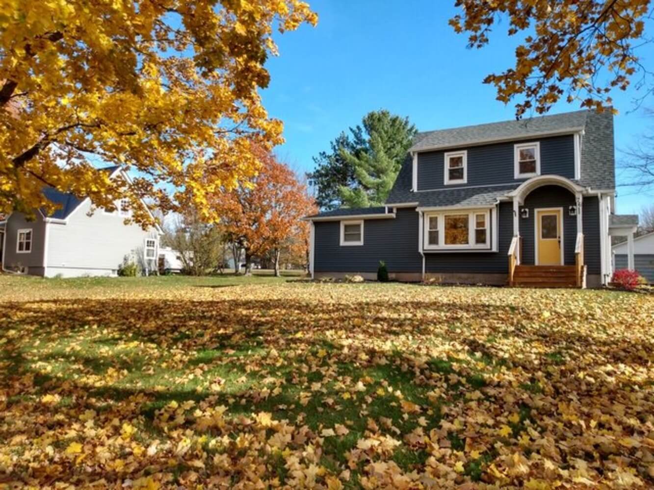 A two-story home with dark blue siding surrounded by fall foliage, illustrating the seasonal window for exterior house painting.